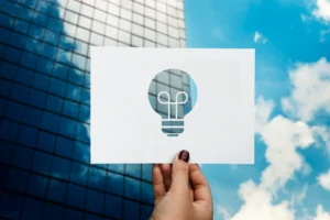 Imagem de uma mão segurando uma cartolina com símbolo de uma lâmpada, representando inovação, com prédio de escritórios ao fundo e céu azul, destacando o papel da inovação na retomada do crescimento econômico.