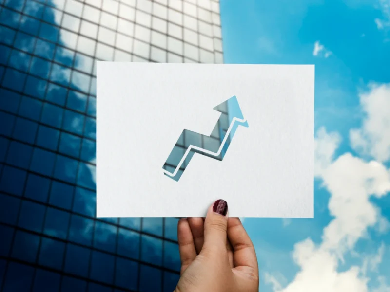 Imagem de uma mão segurando um cartaz com uma seta ascendente representando crescimento econômico, com edifícios modernos ao fundo, relacionado ao desenvolvimento industrial do Brasil para 2026.