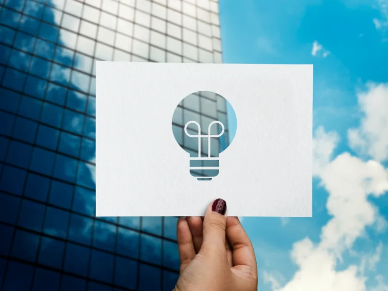 Imagem de uma mão segurando uma cartolina com símbolo de uma lâmpada, representando inovação, com prédio de escritórios ao fundo e céu azul, destacando o papel da inovação na retomada do crescimento econômico.