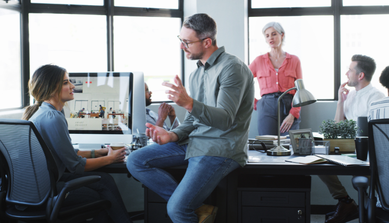 Equipe de profissionais discutindo ideias em um escritório moderno, com computadores e colegas trabalhando ao fundo.